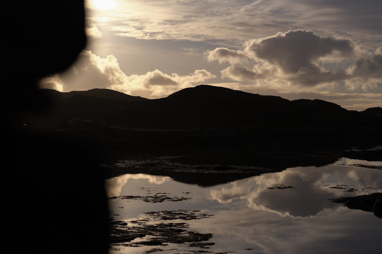 Silhouetted hills and clouds reflected in still water on the Isle of Harris, framed by dark rock